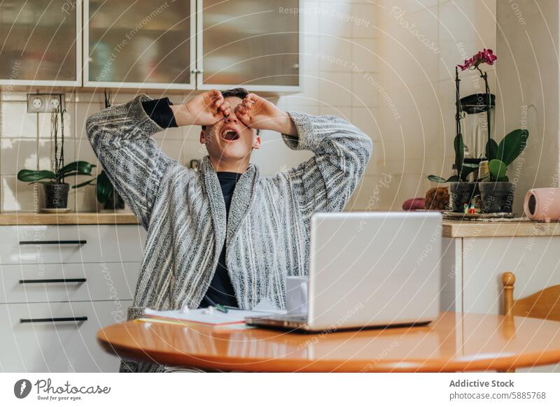 Man stressed in home office with hands over eyes man table laptop robe distress frustrated kitchen indoor orchid plant pot sitting emotion expression anxiety