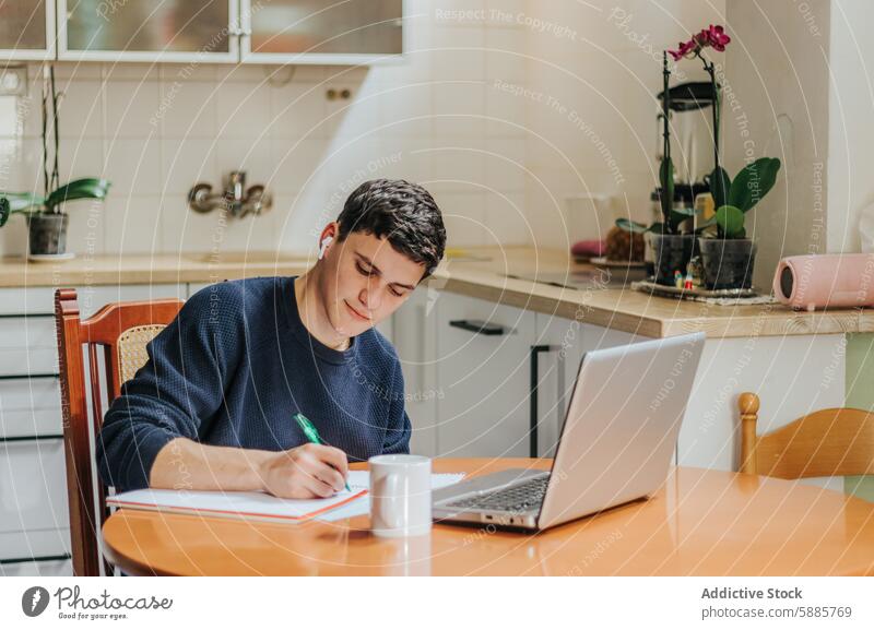 Young man studying on laptop at kitchen table young home environment productive serene session work note taking sit focus diligent warm indoor gadgets student