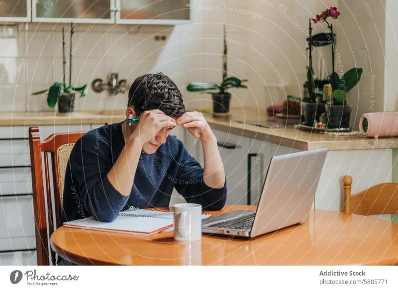 Young man feeling stressed while working from home laptop kitchen young adult table chair frustration technology notebook cup sweater glasses indoor daytime