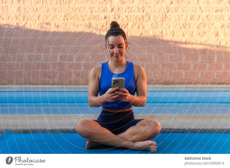 Young woman using a mobile phone on a yoga mat next to a serene swimming pool at sunset smartphone fitness health serenity wellness meditation exercise outdoor