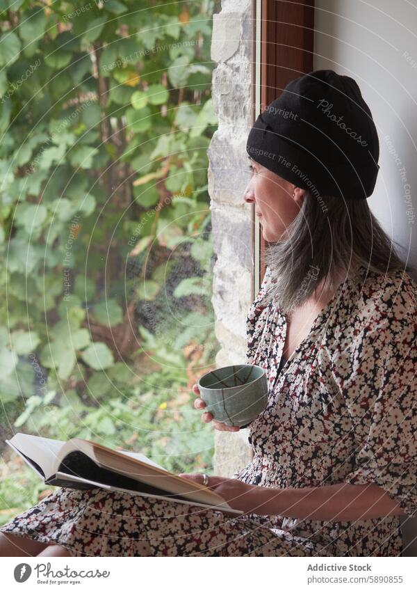 Woman enjoys tea and book by window in autumn woman reading floral dress beret indoors serene peaceful calm leisure relaxation foliage greenery cup holding