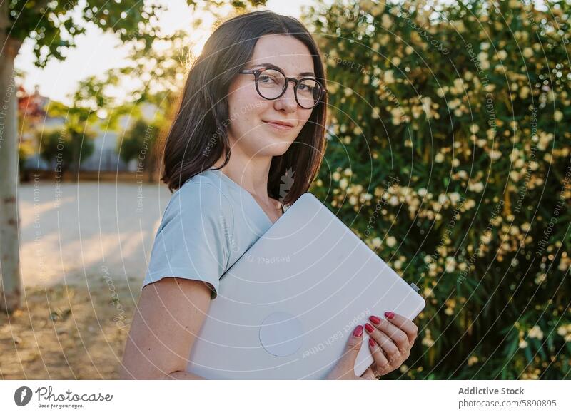 Young woman holding laptop outdoors in a sunny setting tree sunlight glasses smile summer technology gadget device young female adult looking at camera nature