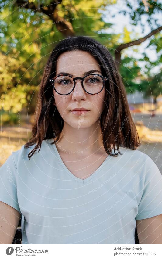 Young woman with glasses looking at camera in a park young portrait caucasian light blue t-shirt neutral expression tree background female person freckles