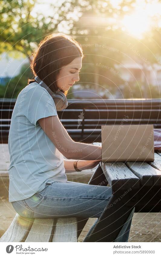 Young woman writing in notebook on park bench headphone concentration backlit sitting looking down write outdoor summer leisure study pen table thoughtful