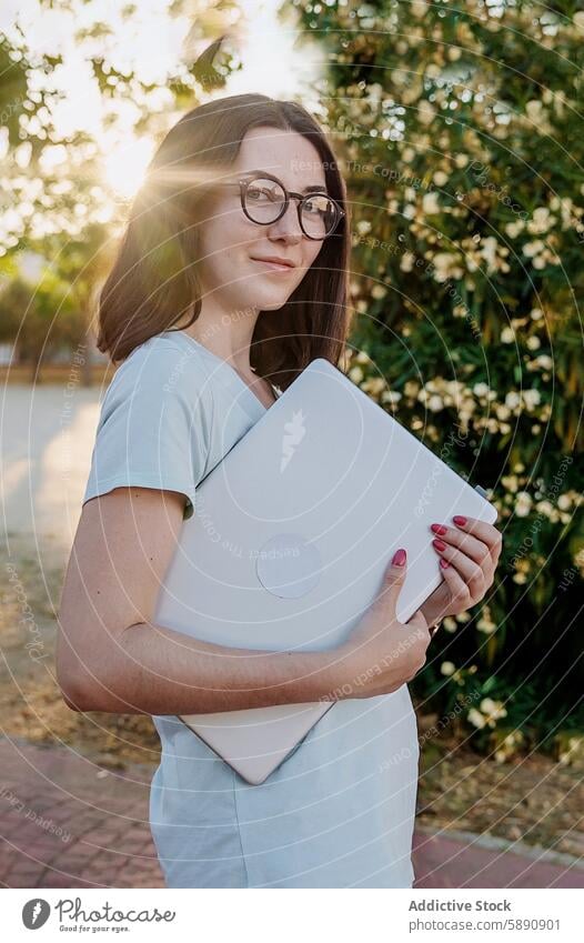 Young woman with glasses holding a laptop outdoors garden sunlight technology gadget device female young embrace brown hair casual summer day nature peaceful