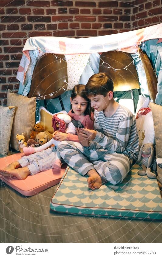 Happy little girl embracing rag doll while looking boy playing video game with phone sitting on tent happy telephone mobile cell children having fun using