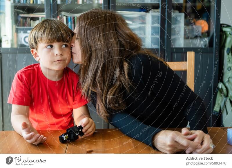 Loving mom kissing cute son on cheek while sitting at table mother love play toy bonding affection relationship motherhood childhood playful at home adorable