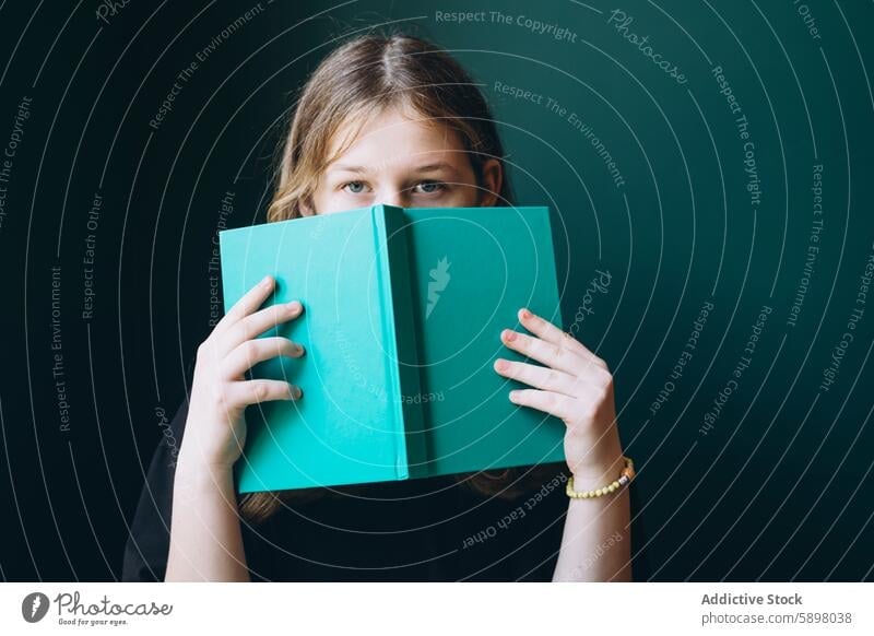 Young girl peering over a green book, looking at camera student education back to school study learning reader youth eye contact schoolgirl literature knowledge