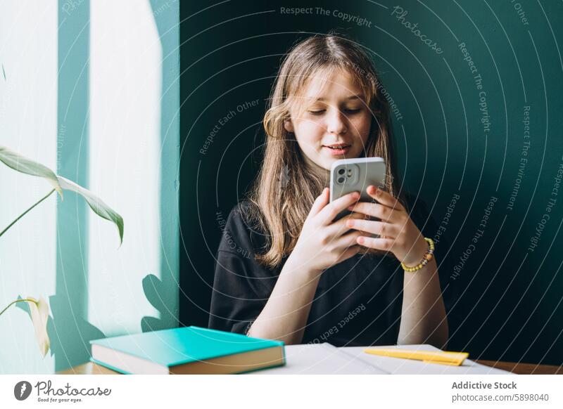 Teenage girl using smartphone at her study desk teenager studying book plant education school technology gadget mobile phone looking down indoor learning