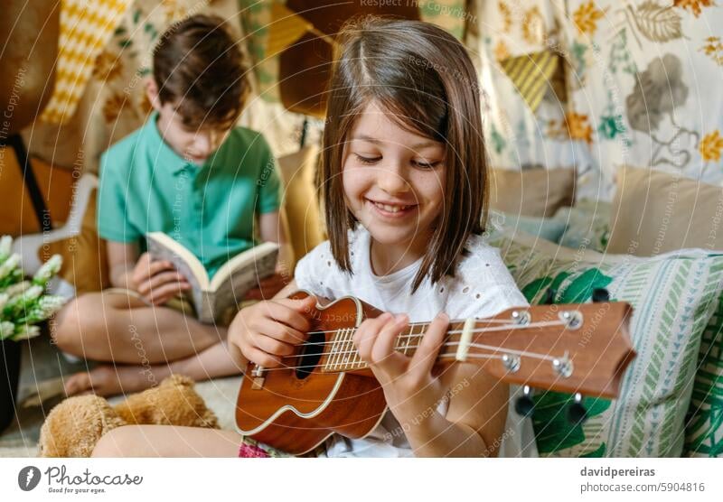 Happy smiling little girl playing ukulele while boy reading book on handmade shelter tent happy smile guitar teepee homemade portrait children camping having