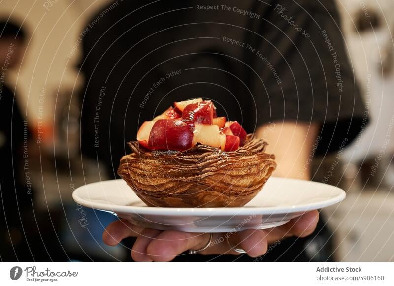 Close-up of pastry topped with fresh fruit held by waiter unrecognizable anonymous faceless serving white plate blurred background dessert close-up food gourmet