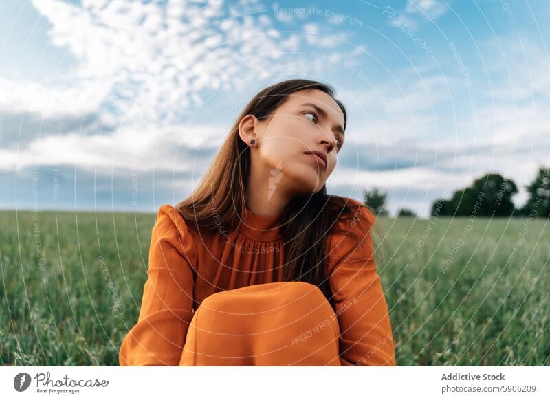 Young brunette woman looking away in a grassy field sky clouds serene thoughtful outdoors nature green orange blouse vibrant peaceful young female relax leisure