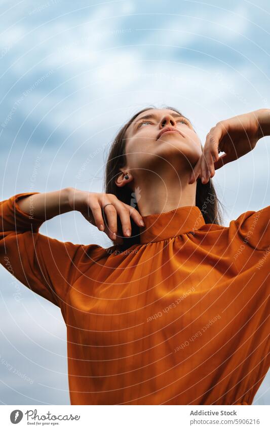 Serene brunette woman looking up in an orange dress outdoors serene touching neck cloudy sky contemplative thoughtful gentle fashion elegance beauty stylish