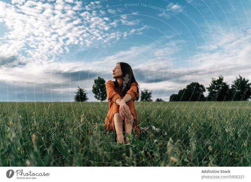 Serene woman in orange dress enjoying nature in a field serene brunette looking away contemplative outdoors green sky cloudy elegant sit lush landscape summer