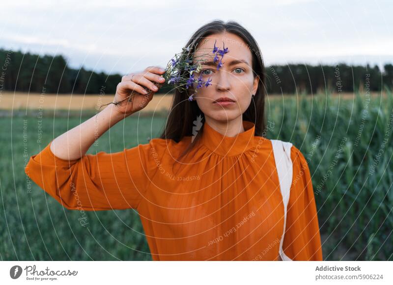 Brunette woman holding flowers in a field, looking at camera brunette gaze serene orange blouse green wildflower outdoor nature expression summer fashion style