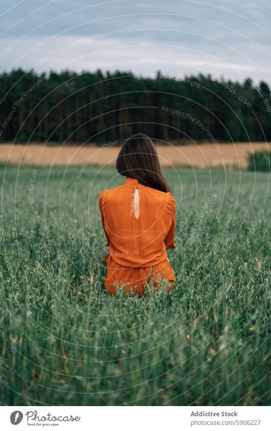 Brunette woman in orange dress standing in a green field brunette alone serene nature outdoors vibrant distant forest cloudy sky summer countryside solitude