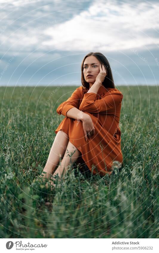 Thoughtful brunette woman sitting in a green field orange dress grass pensive thoughtful contemplative nature outdoor summer leisure alone calm young female