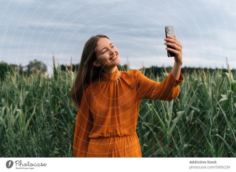 Young brunette woman taking selfie in a green cornfield smartphone orange dress joy outdoor mobile phone nature agriculture summer casual leisure device gadget