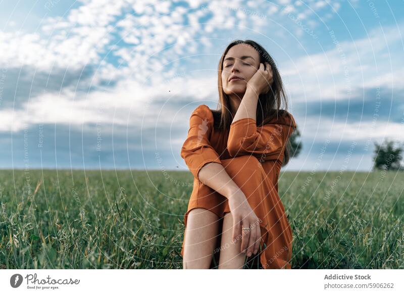 Serene woman in orange dress enjoying nature in a field serene brunette eyes closed contemplative outdoors green sky cloudy elegant sit lush landscape summer