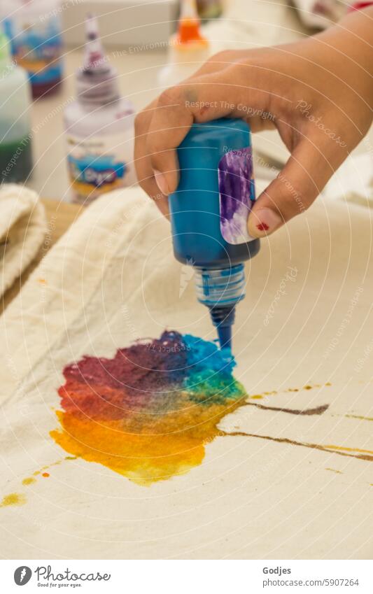 Creative time - colorful cloud is dripped onto the fabric with fabric paint, hand holds bottle with the paint, other painting utensils can be seen in the background