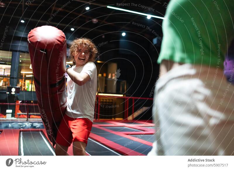 Joyful man playing with punching bag at jumping club indoor laughter fun curly hair energetic playful red white shirt red shorts male active lifestyle joy