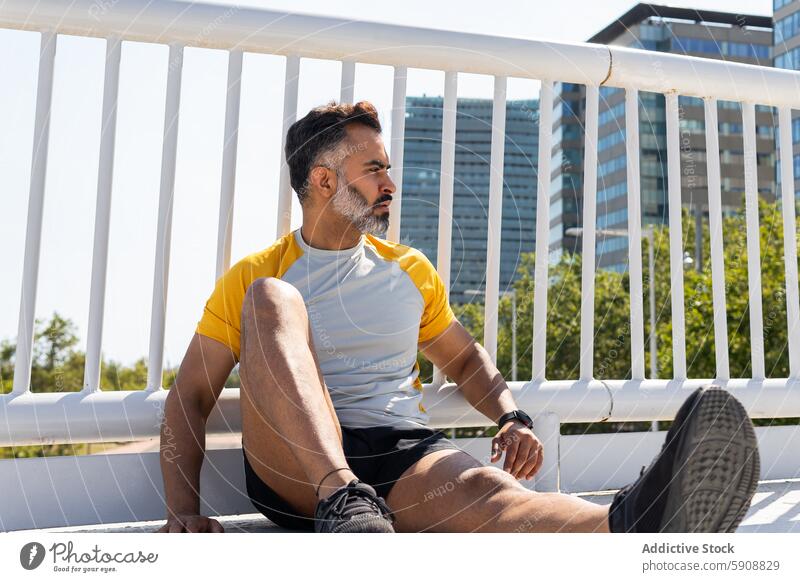 Man resting during summer workout in urban setting indian sport man sitting looking away city building sportswear t-shirt shorts sneakers sunny outdoors fitness