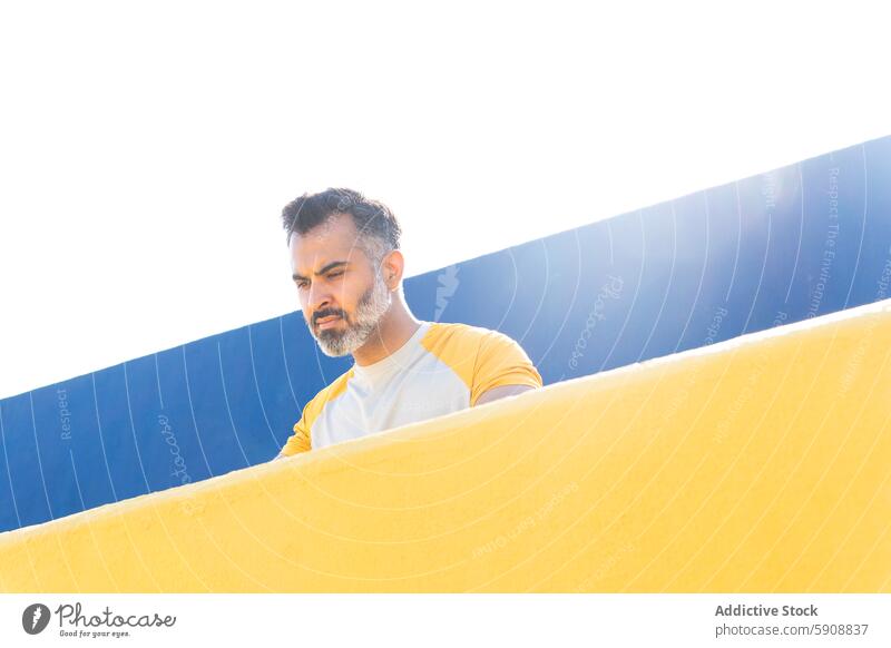 Indian man in sporty attire leaning on yellow wall indian summer blue sky clear sunny outdoor casual relaxed thoughtful gaze looking down active fitness