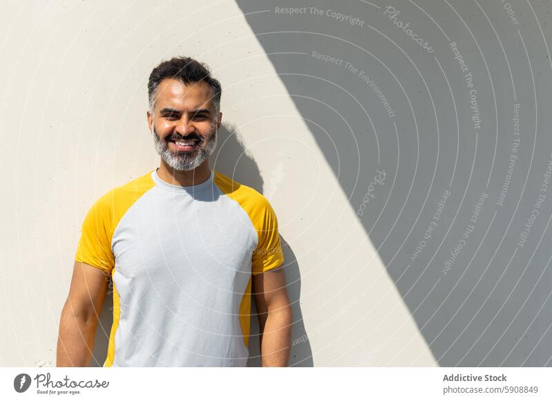 Smiling Indian man in casual summer attire standing against a wall indian smile yellow white t-shirt cheerful shadow sunlight day outdoor warm happy facial hair