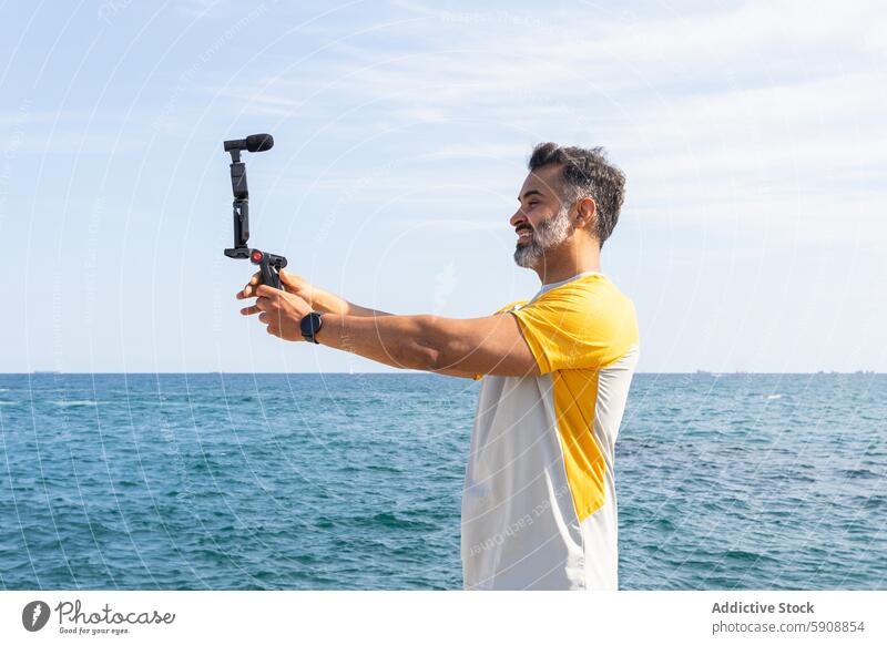 Man recording video with gimbal at the seaside indian man sport summer technology device gadget ocean water blue sky outdoor looking at screen leisure