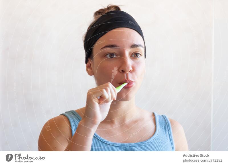 Woman brushing teeth in a simple bathroom setting woman hygiene self-care routine oral care toothbrush headband tank top health toothpaste personal wellness