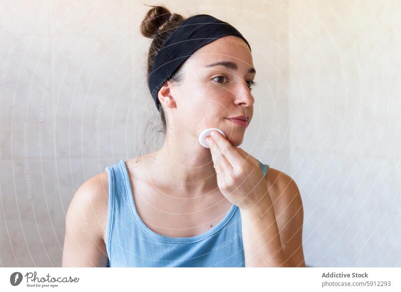 Woman practicing a skincare routine in a bright bathroom woman self-care morning cleansing face cotton pad casual tank top headband serene gentle cleanse