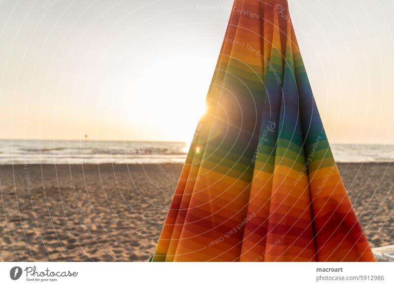 Colorful striped, closed parasol in close-up on the beach at sunset variegated Striped Sunshade Closed too Sand Beach Sandy beach Ocean Water Sunbeam Sunset