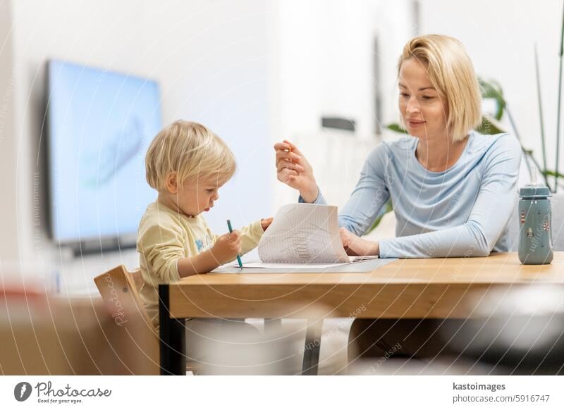 Caring young Caucasian mother and small son drawing painting in notebook at home together. Loving mom or nanny having fun learning and playing with her little 1,5 year old infant baby boy child.