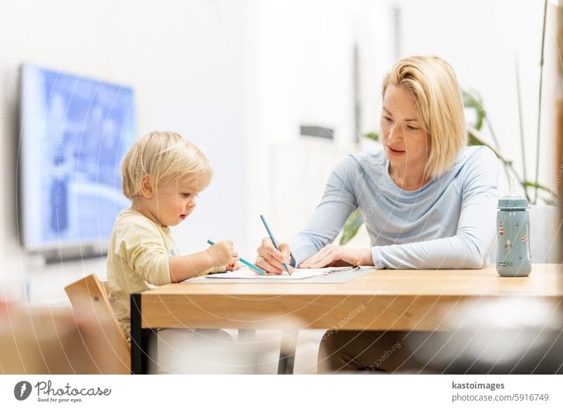 Caring young Caucasian mother and small son drawing painting in notebook at home together. Loving mom or nanny having fun learning and playing with her little 1,5 year old infant baby boy child.