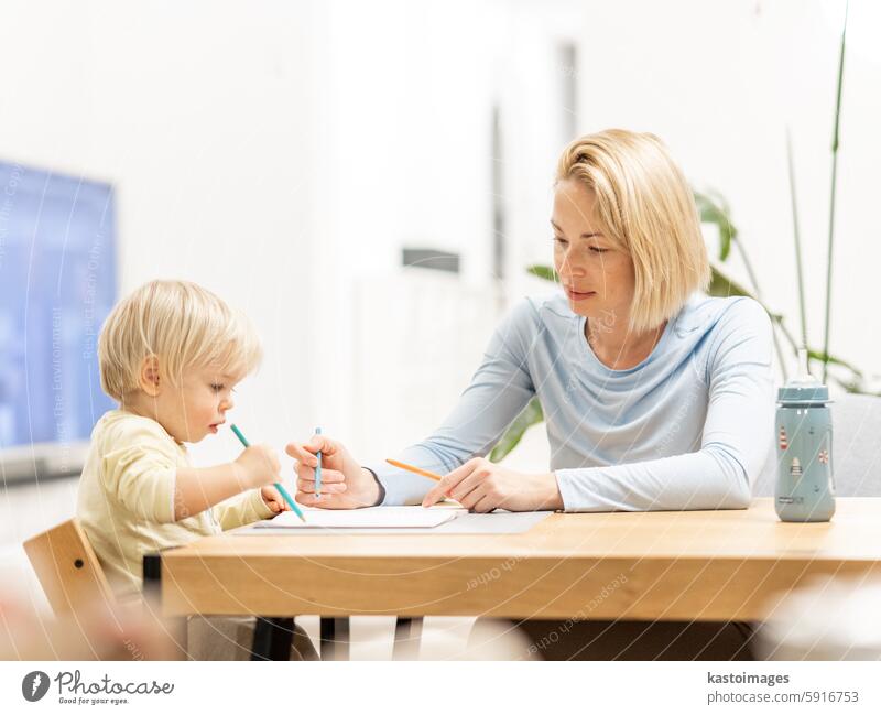 Caring young Caucasian mother and small son drawing painting in notebook at home together. Loving mom or nanny having fun learning and playing with her little 1,5 year old infant baby boy child.