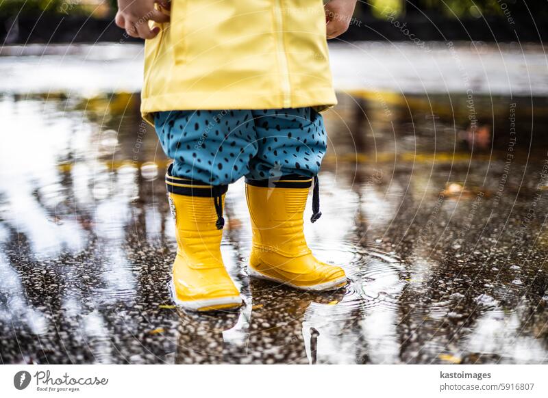 Small infant boy wearing yellow rubber boots and yellow waterproof raincoat standing in puddle on a overcast rainy day. Child in the rain. child fun weather