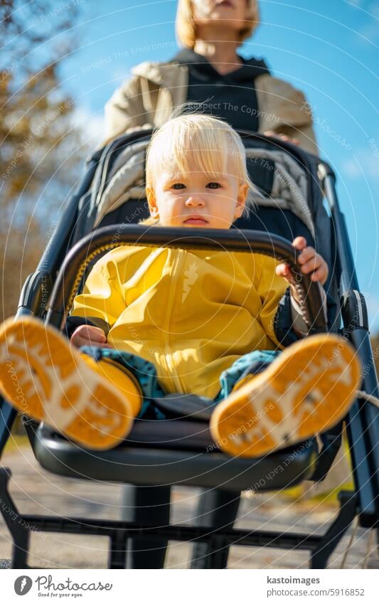 Little toddler child wearing yellow rain coat and rain boots being pushed in stroller by her mother in city park on a sunny autumn day after the rains were gone.