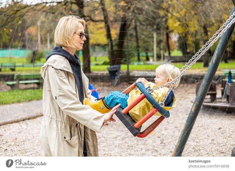 Mother pushing her infant baby boy child wearing yellow rain boots and cape on swing on playground outdoors on cold rainy overcast autumn day in Ljubljana, Slovenia