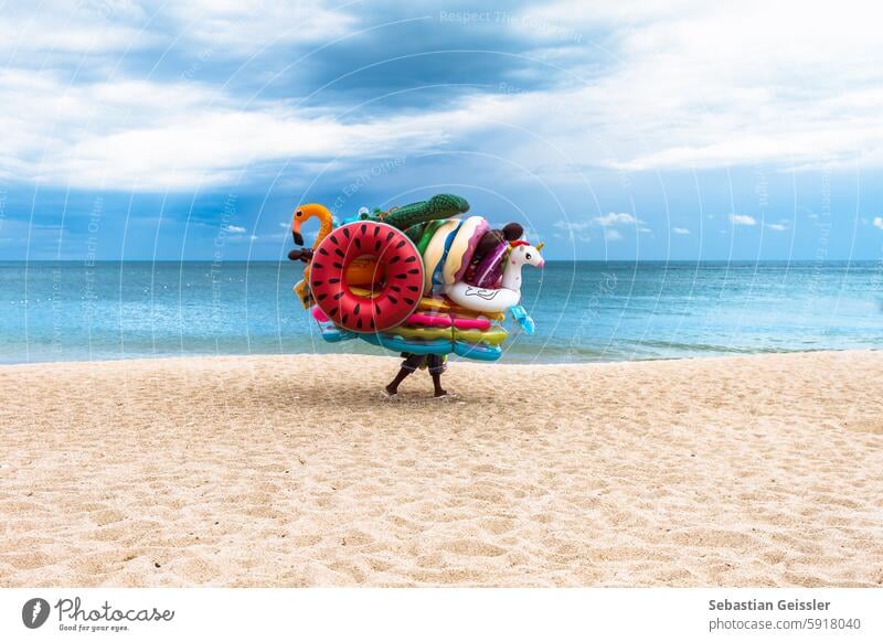 The picture shows a scene on a sandy beach with a calm sea and a cloudy sky in the background. In the foreground, a person is carrying a large number of inflatable swimming toys and swimming rings.