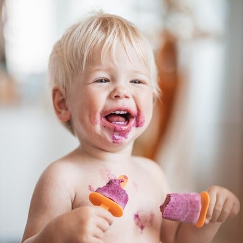 Happy adorable infant baby boy child smiling while eating two frozen fruit popsicle ice creams in simmer. kid food summer fun childhood snack little cute