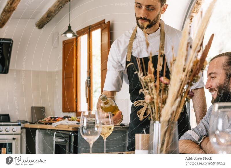 Private chef serving wine to a man at a kitchen gathering private chef white wine smiling guest pouring counter warm inviting rustic atmosphere looking down