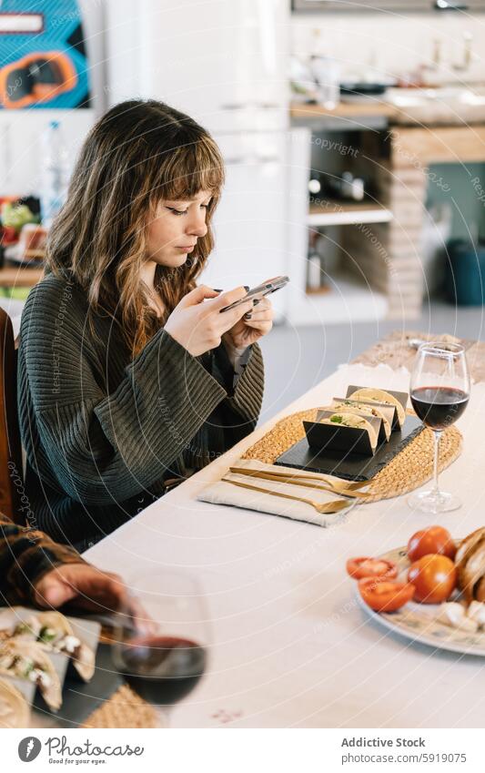 Young woman using phone during a meal with friends smartphone dining table dishes wine glass red wine cozy home social food technology indoor natural light