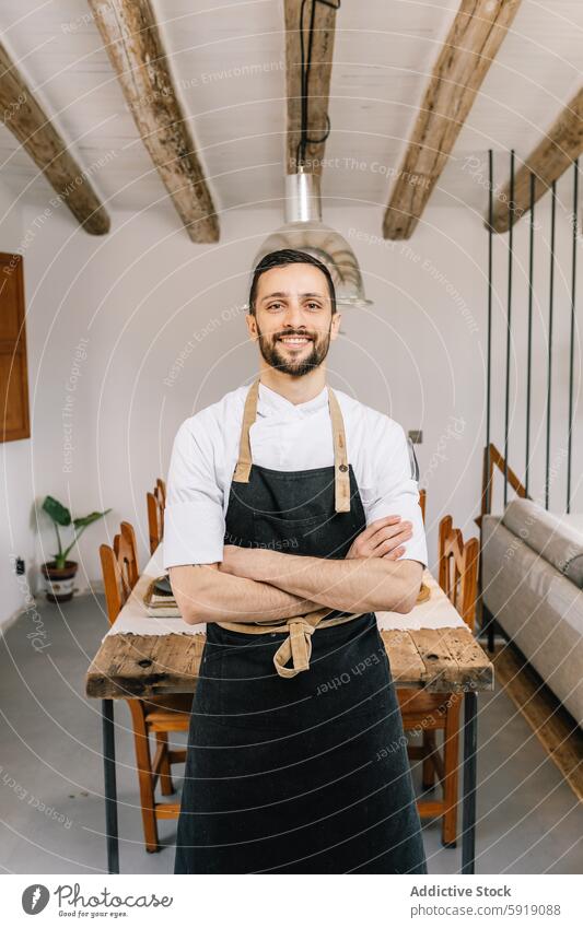 Confident male chef in rustic kitchen smiling at the camera apron professional arms crossed confident indoor wooden table cozy warmth white shirt beams ceiling