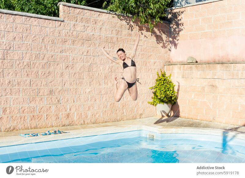 Young woman joyfully jumping into a swimming pool young sunlight summer fun leisure backyard outdoor activity swimwear happiness energetic brick wall greenery