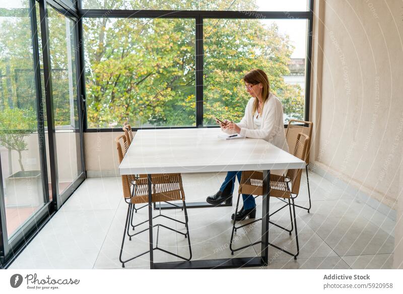 Mid-adult woman using a smartphone at a modern outdoor cafe table tranquil park view seated browsing chic white well-lit background mid-adult chair window glass