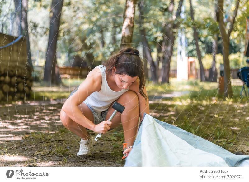 Focused young woman holding a hammer while staking a tent into the ground against a wooded campsite casual attire involvement simplicity joy nature connecting