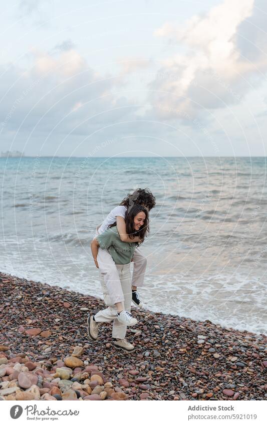 Smiling woman carrying homosexual beloved piggyback on ocean beach girlfriend lesbian having fun carefree relationship embrace coast women twilight sea pebble