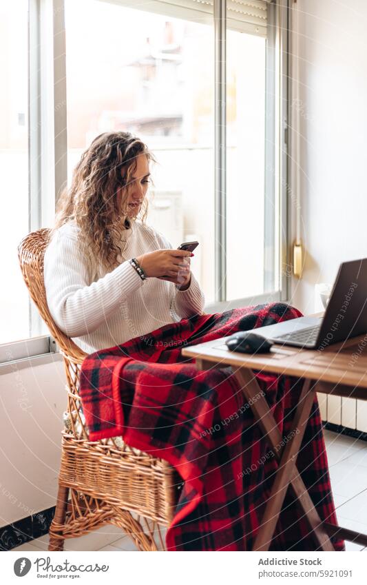 Young woman browsing smartphone while sitting at table with laptop using mobile freelance work at home surfing remote connection online internet cellphone