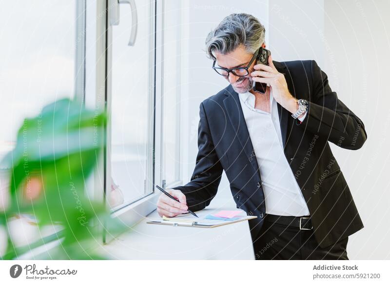 A focused executive multitasks, managing his workload with a phone tucked between shoulder and ear, pen in hand, near a bright window call office happy business