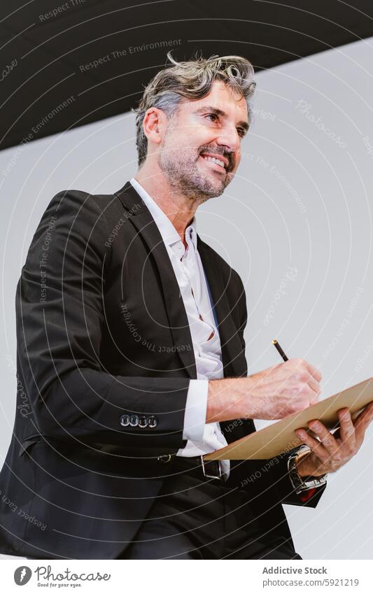 From below of cheerful mature executive in a sleek suit holds a clipboard, reviewing contents with a pleased expression in a stylish office man smiling business